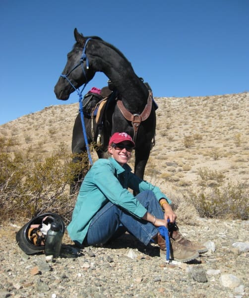 A person in a green shirt and red cap sits on rocky ground beside a black horse with a saddle and blue reins in a desert landscape under a clear blue sky.