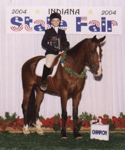 Person in riding attire holding a plaque while sitting on a horse decorated with a green garland. A "Champion" sign is in front and "Indiana State Fair 2004" banner is in the background.