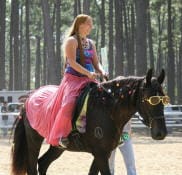 A woman dressed in a colorful outfit rides a horse decorated with fairy lights and wearing sunglasses, in an outdoor area surrounded by tall trees.