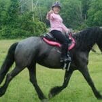 A person wearing a pink shirt and helmet smiles and gives a thumbs up while riding a dark gray horse in a grassy field.