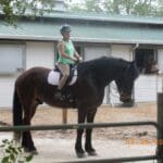 A person in a green top and helmet sits on a brown horse in front of a stable. Another horse stands with its head out of the stable window. The date on the photo reads 04/26/2009.