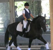 A person rides a black horse in an indoor equestrian facility, both dressed in a formal riding attire.