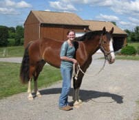 A person stands outside on a gravel area beside a brown horse, holding its lead rope. There is a barn in the background with a partly cloudy sky.