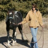 A person in a tan jacket and blue jeans is leading a dark-colored horse with a white marking on its face using a rope, walking outdoors on a dirt path with trees in the background.