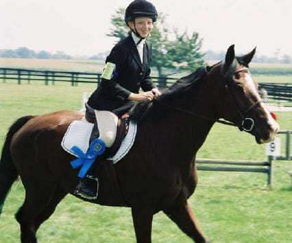 A rider wearing a helmet and riding attire sits on a horse in an open grassy field, holding a blue ribbon.