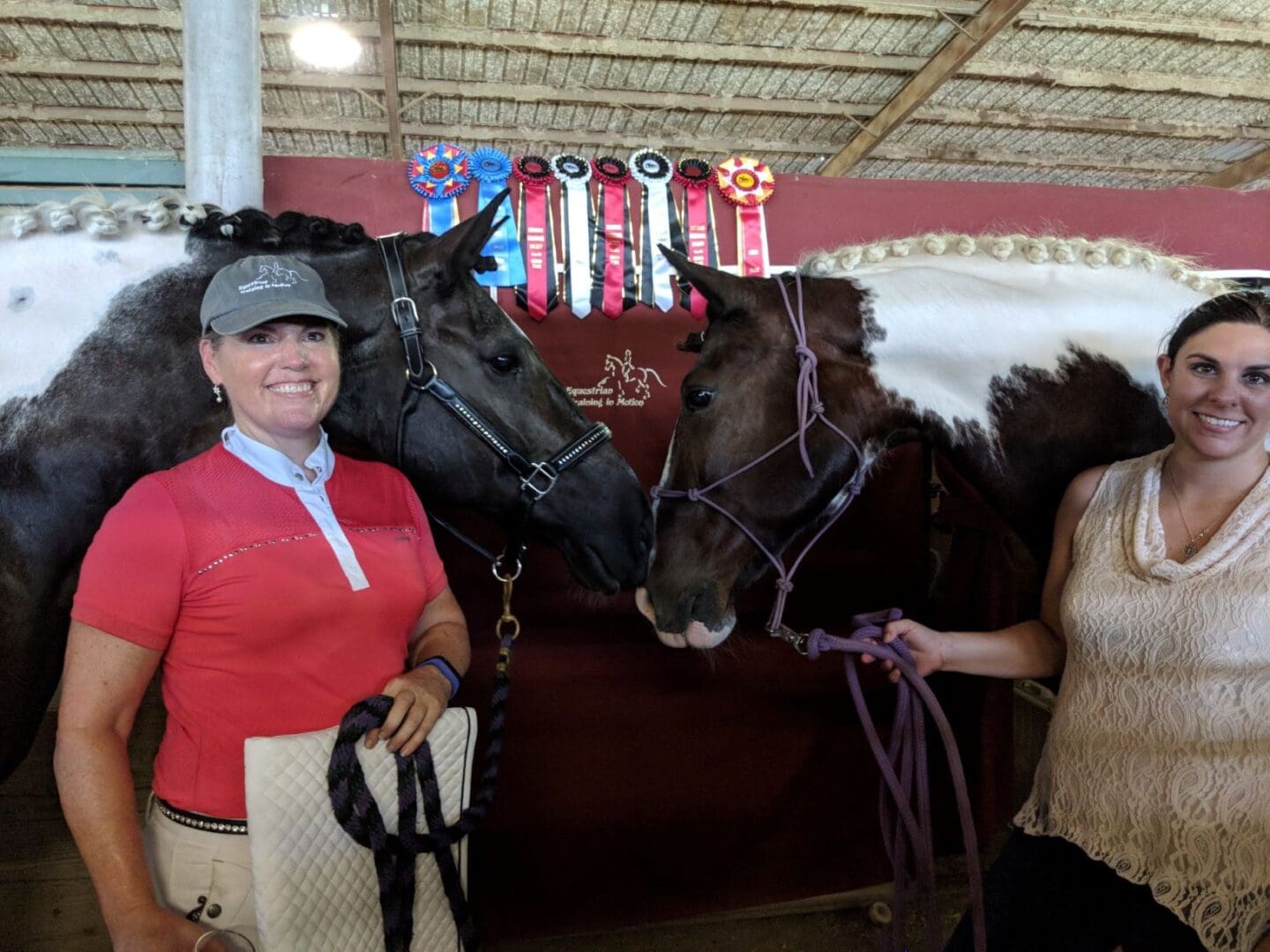 Two women stand with horses in front of a wall displaying numerous ribbons. One woman, in a red shirt and cap, holds a blanket, while the other, in a sleeveless top, holds a horse's reins.