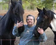 A person stands outdoors, touching the heads of two horses on either side while smiling at the camera.