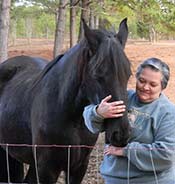 A person with short gray hair pets a black horse over a fence in a wooded area.