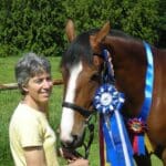 A woman stands beside a horse adorned with multiple blue and red award ribbons around its neck in a grassy area with trees in the background.