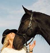 A woman wearing a black hat and white shirt smiles at a black horse, which nuzzles its head close to her.