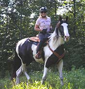 A person wearing a helmet and sunglasses rides a black and white horse through a wooded area.