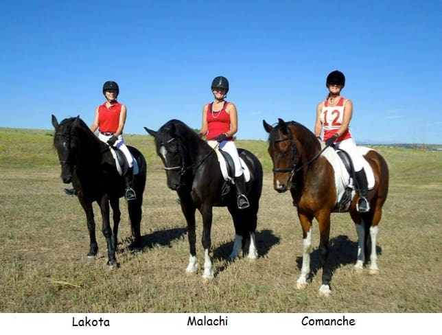 Three equestrians in matching red tops and riding helmets sit on horses in an open field. Below each rider, the names "Lakota," "Malachi," and "Comanche" are labeled corresponding to the horses.