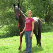 A woman stands on grass holding the lead of a dark-colored horse with a red halter. Trees in the background.