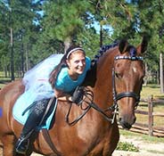 A person in a blue outfit and tiara rides a brown horse with a braided mane in an outdoor setting surrounded by trees.