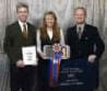 Three people stand together holding an award ribbon, a plaque, and a banner. The banner reads "Best in Show, Platinum Award, 2016." They are dressed in formal attire.