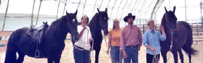 Four people standing side by side, each holding the reins of their respective horses, inside a large indoor riding arena.