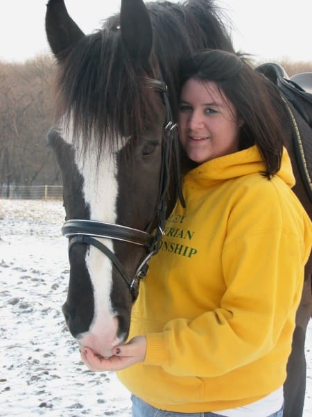 A woman in a yellow hoodie stands next to a horse with a white blaze, petting its nose in a snowy outdoor setting.