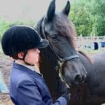 A person wearing a helmet and blazer stands next to a black horse, holding its bridle.