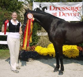 A person stands holding ribbons next to a dark-colored horse. Behind them, a sign reads "Dressage at Devon." Yellow flowers are placed in the background.