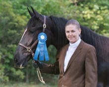 Person in a brown jacket stands next to a dark horse adorned with a blue ribbon in an outdoor setting.