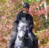 A person wearing a helmet and dark clothing is riding a gray horse through a wooded area with autumn foliage.