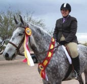 A person in riding gear sits atop a gray horse adorned with a ribbon, outdoors with trees and a cloudy sky in the background.