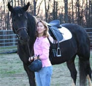 A person wearing a pink shirt holds a black horse by the reins, standing in a grassy area with a wooded background and a metal fence. The person is also holding a riding helmet in one hand.