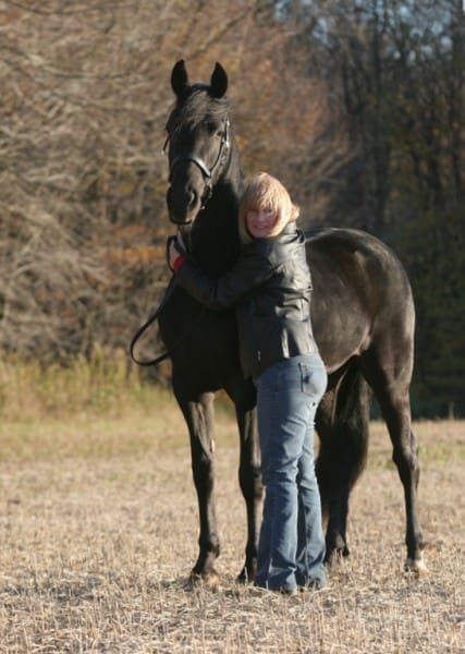 A person wearing a jacket and jeans hugs a tall, black horse in a field with autumn trees in the background.